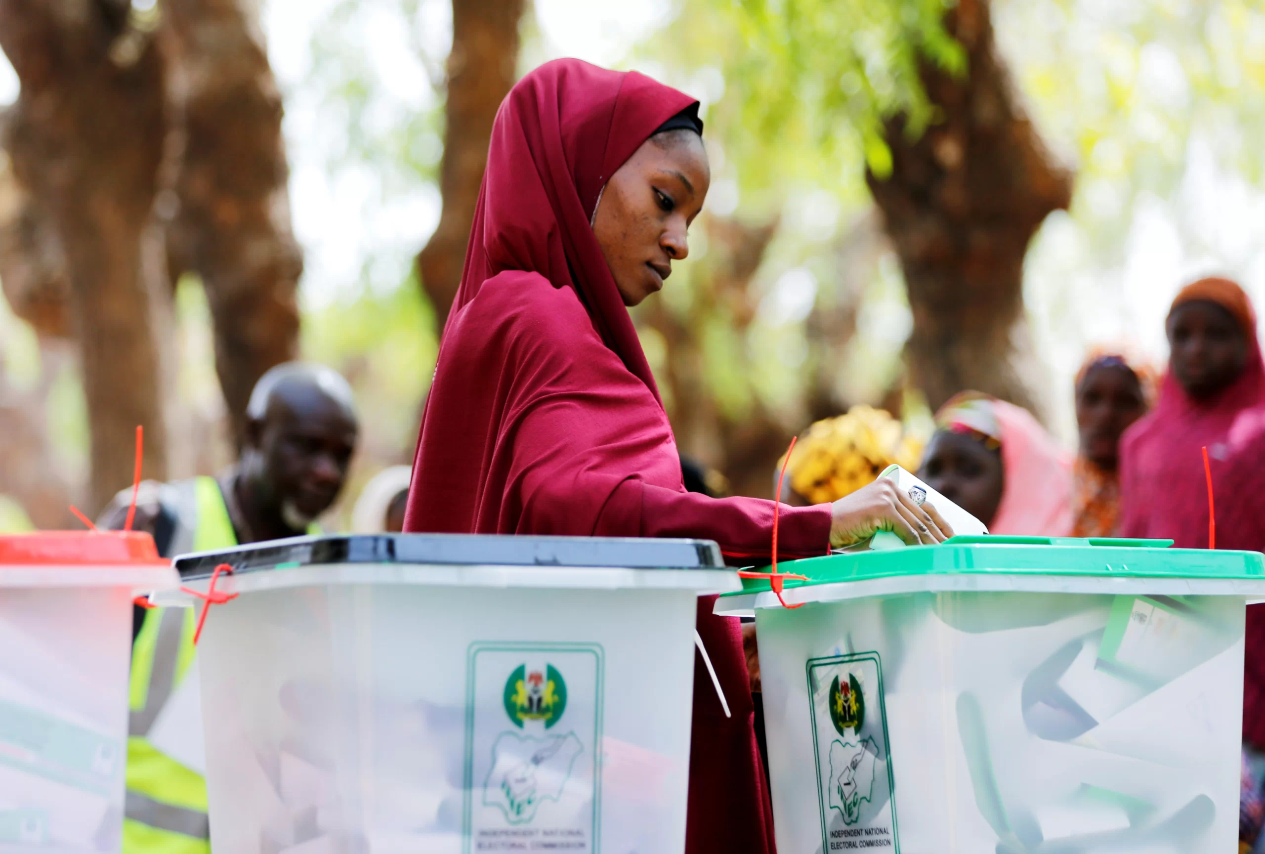A Nigerian woman in a red hijab casting her ballot into a transparent INEC ballot box